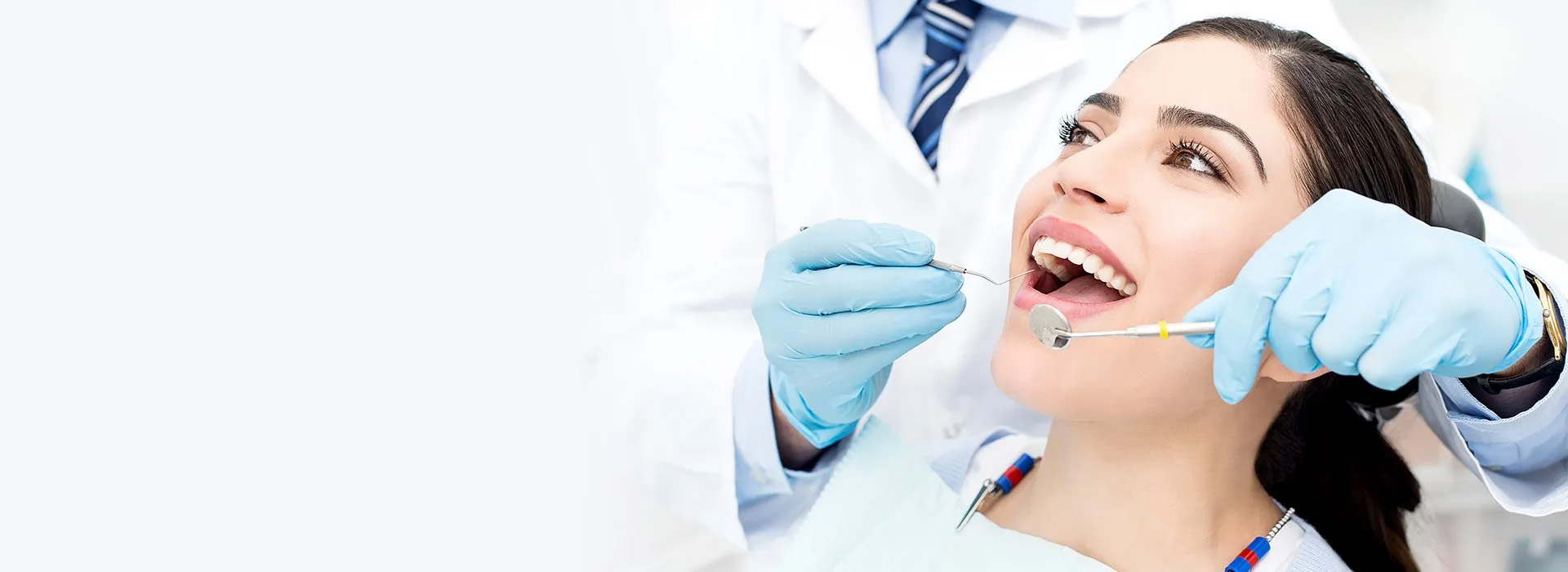 A woman is smiling in a dental chair while a dentist examines her teeth. The dentist is wearing blue gloves and holding dental tools, conveying a professional, calm atmosphere.
